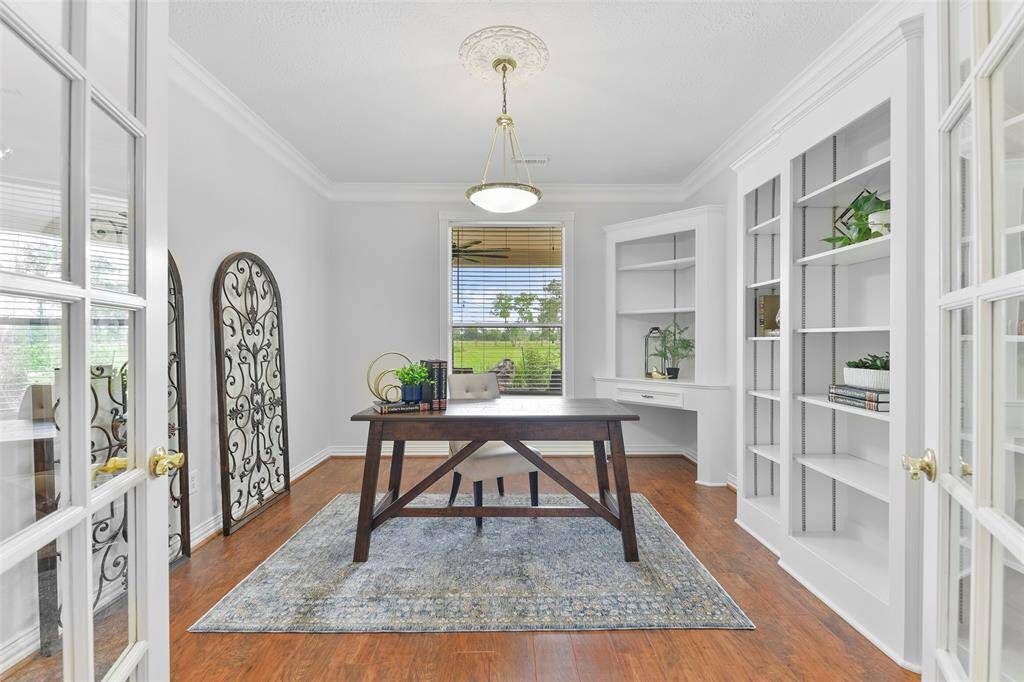 A pair of French doors create privacy for this handsomely appointed home office that features luxury vinyl plank flooring, a wall of adjustable built-in shelving, double crown molding, fresh paint, a pendant light with a ceiling medallion, and a rear-facing window with 2-inch blinds.