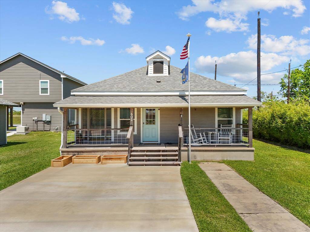 Charming single-story home with a welcoming front porch featuring rocking chairs. The house has a neatly manicured lawn and a spacious driveway. It has a classic design with a gabled roof and a flagpole in front, adding a touch of character.
