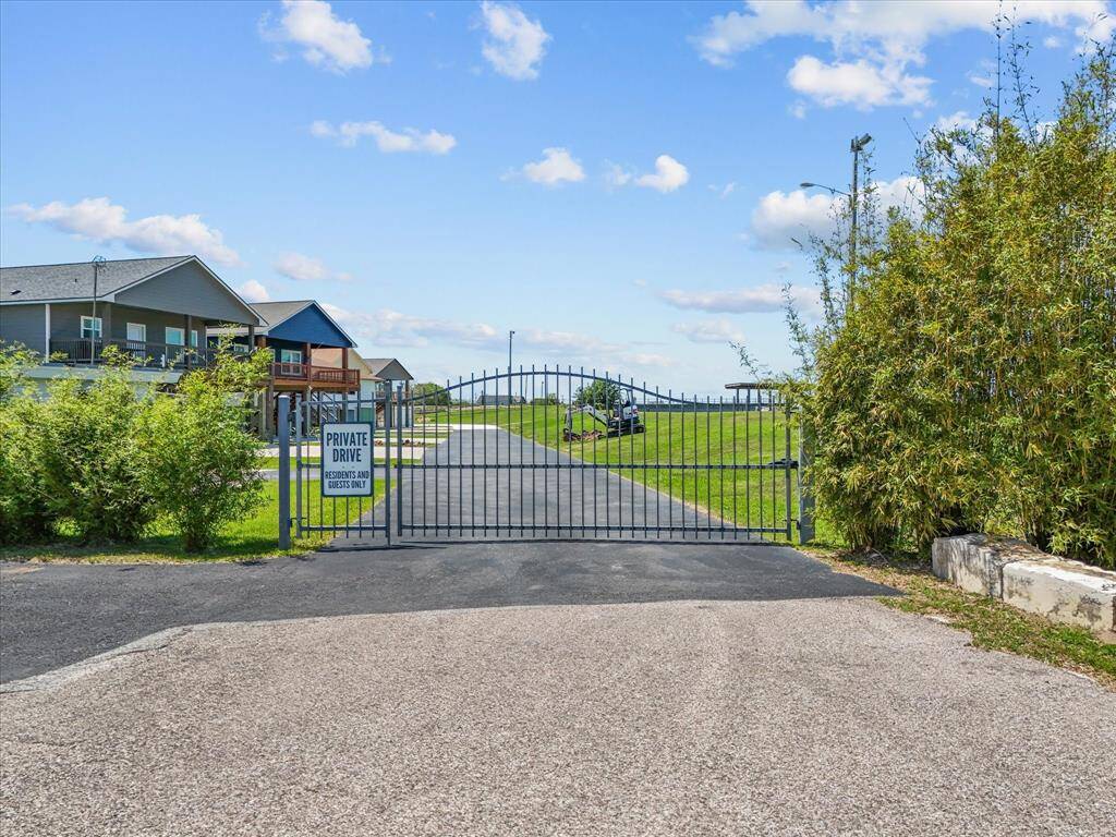 Gated entrance to a private residential area, offering security and exclusivity. The drive is bordered by greenery, leading to modern homes with large porches. Ideal for buyers seeking privacy in a community setting.