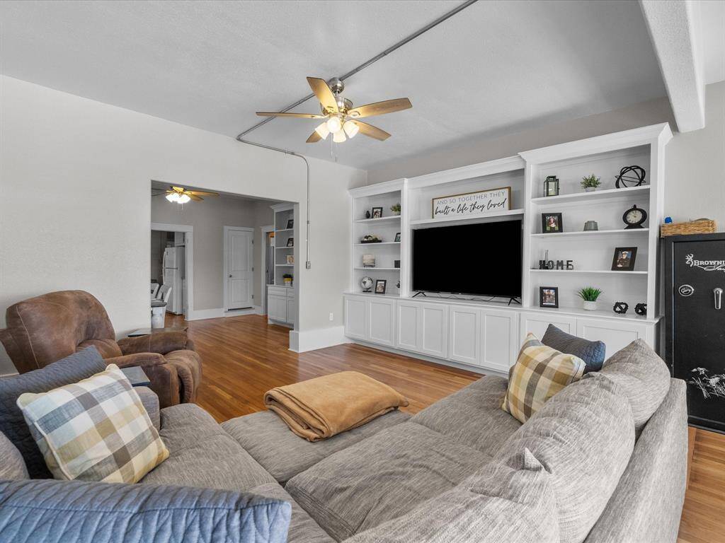 Spacious living room with hardwood floors, a ceiling fan, and built-in shelving around a large TV. Cozy seating area with a neutral color palette and decorative accents. Open doorway leads to additional rooms.