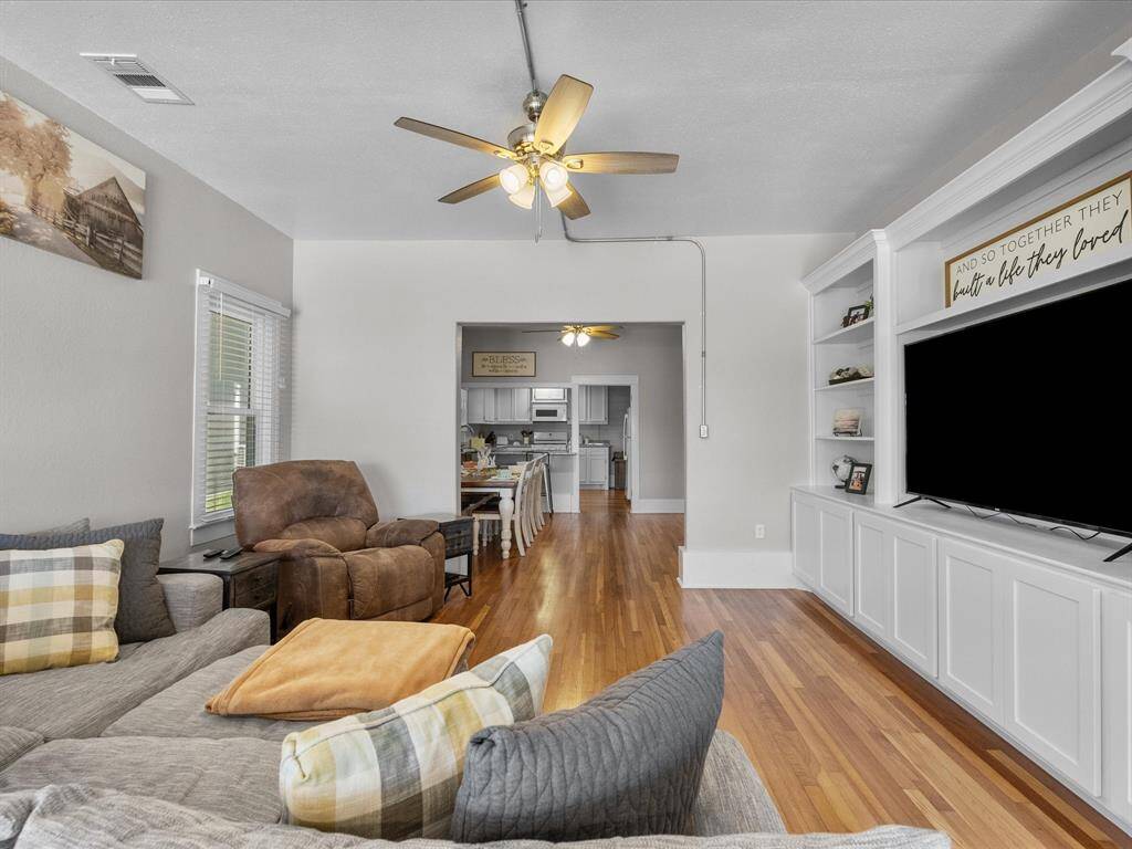 This inviting living room features hardwood flooring, a ceiling fan, built-in shelving with a large TV, and cozy seating. It opens up to a dining area and kitchen, creating a seamless flow for entertaining.