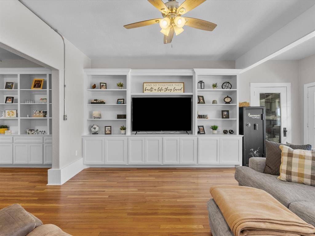 This inviting living room features built-in white shelving and cabinets, a central TV, and warm wood flooring. A ceiling fan adds comfort, while cozy seating and decorative accents create a welcoming atmosphere.