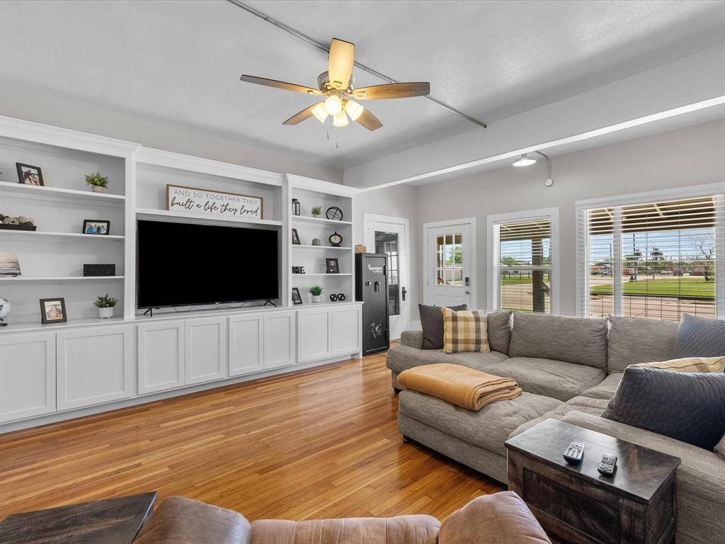 This inviting living room features warm hardwood floors, built-in white shelving, and a large TV. It has a cozy sectional sofa, ceiling fan, and ample natural light from windows and a glass door leading outside. The space is perfect for relaxation and entertainment.
