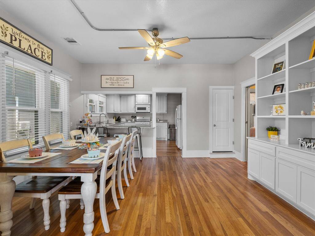 This photo showcases a bright dining area with hardwood floors and a ceiling fan, leading into a white, modern kitchen. There's ample natural light from large windows, and built-in shelving adds charm and storage. A long dining table offers plenty of seating for gatherings.