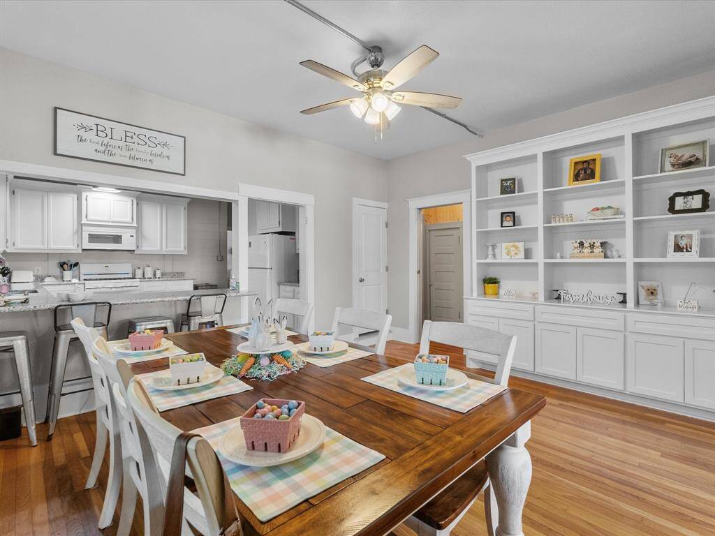 Bright dining area featuring a wooden table set for six, built-in white shelving, and an open view into a modern kitchen. The space is accented with a ceiling fan and hardwood floors, offering a cozy and welcoming atmosphere.