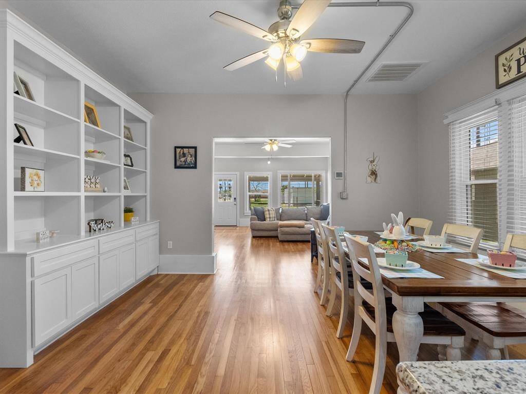 This inviting space features an open floor plan with warm wood flooring, a spacious dining area, and built-in shelving for storage. Natural light fills the room, highlighting the cozy living area in the background, perfect for family gatherings.