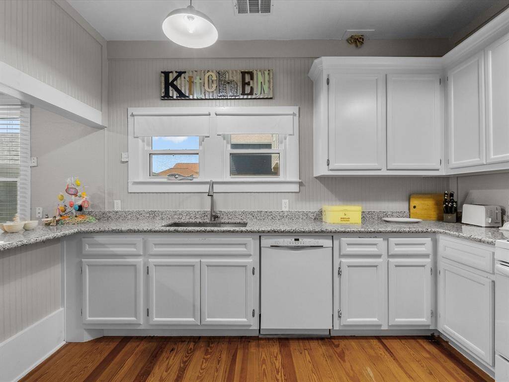 This kitchen features bright white cabinetry, granite countertops, and hardwood floors. It includes a dishwasher and ample natural light from the window above the sink. The space is accented with a decorative "Kitchen" sign and modern lighting.