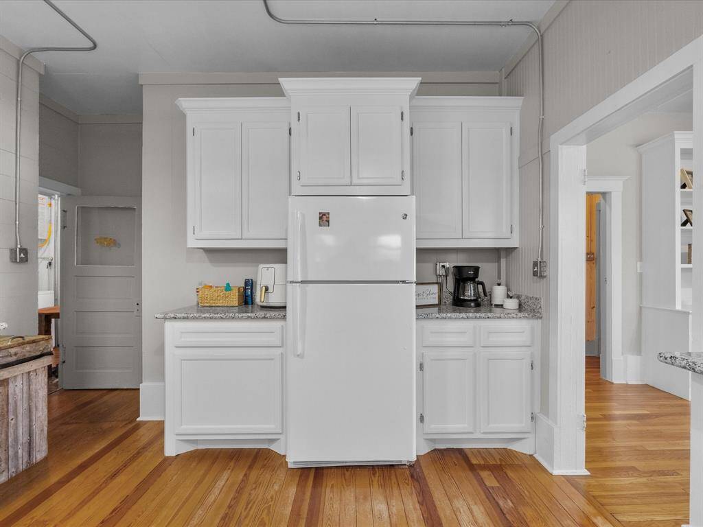 This kitchen features white cabinetry with granite countertops, a central white refrigerator, and natural wood floors, creating a bright and inviting space.