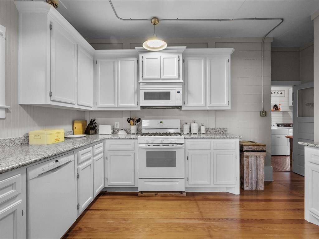 This kitchen features white cabinetry, granite countertops, and a gas stove with an overhead microwave. The wood flooring adds warmth, and there's ample counter space for cooking. The room is well-lit with a classic overhead light fixture.