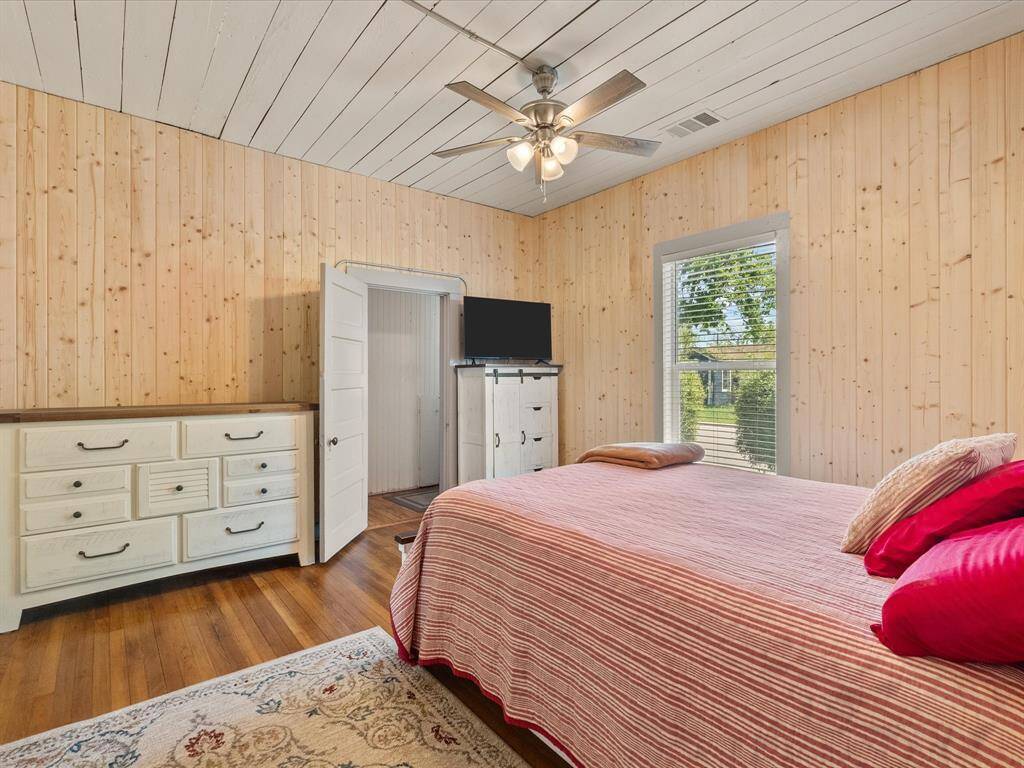Cozy bedroom with warm wood-paneled walls and ceiling. It features a ceiling fan, large window with a view, and two dressers for ample storage. The room has a comfortable bed with colorful bedding and a decorative rug, creating an inviting atmosphere.