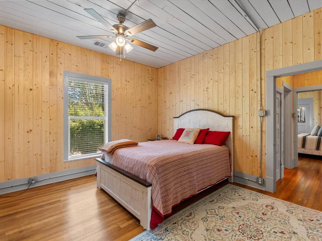 Cozy bedroom with natural wood paneling, hardwood floors, and a ceiling fan. Features a window with garden views and an adjacent room visible through a doorway.