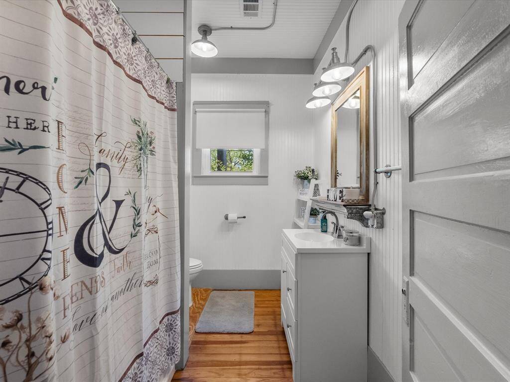 This bathroom features a cozy, farmhouse style with white beadboard walls, a wood-framed mirror, and a vanity sink. It includes a shower with a decorative curtain, a window for natural light, and warm wooden flooring.
