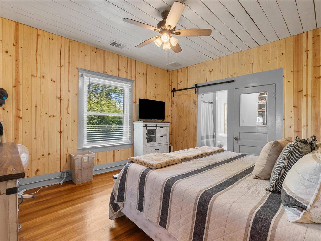 Cozy bedroom with warm wood paneling and hardwood floors, featuring a ceiling fan and natural light from a large window. The room includes a sliding barn door leading to an adjacent bathroom, creating a rustic yet modern feel.