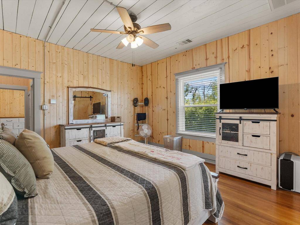 This cozy bedroom features wood paneling, a ceiling fan, and hardwood floors. It includes a window with natural light, a dresser with a mirror, and a TV stand. The room has a warm, rustic feel, perfect for relaxation.