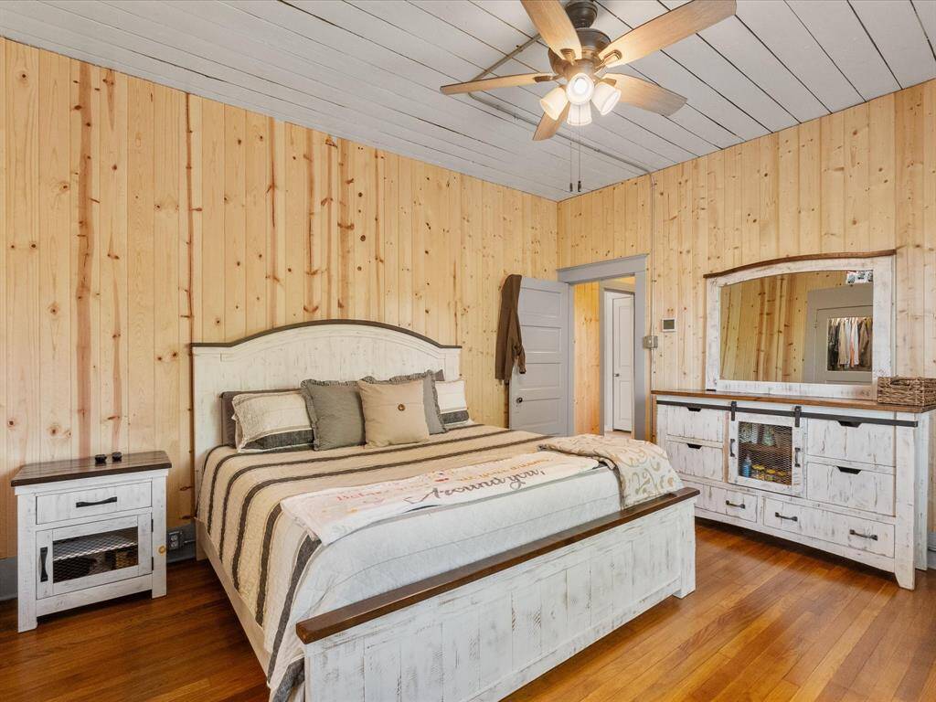 This cozy bedroom features wood-paneled walls and ceiling, creating a rustic feel. It includes a ceiling fan, a large bed with neutral bedding, and matching white distressed furniture, offering ample storage and a warm, inviting ambiance.