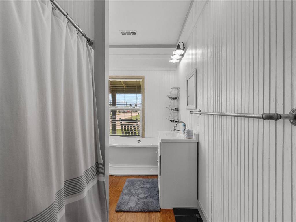 This bathroom features white beadboard walls, a shower with a curtain, a modern vanity with a mirror, and a window with natural light overlooking a tub. A wooden floor and a cozy rug complete the space.