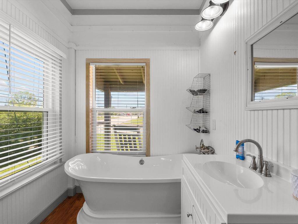 This bathroom features a bright, airy space with a freestanding bathtub next to large windows, offering plenty of natural light. The room has white paneling, a modern vanity with a sleek faucet, and rustic shelving for a charming touch.