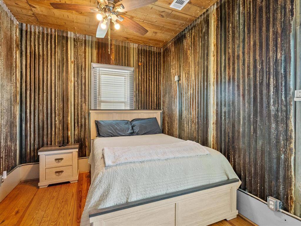 This cozy bedroom features rustic corrugated metal walls and a wooden ceiling with a fan. It includes a comfortable bed and a wooden nightstand, adding to its unique charm.