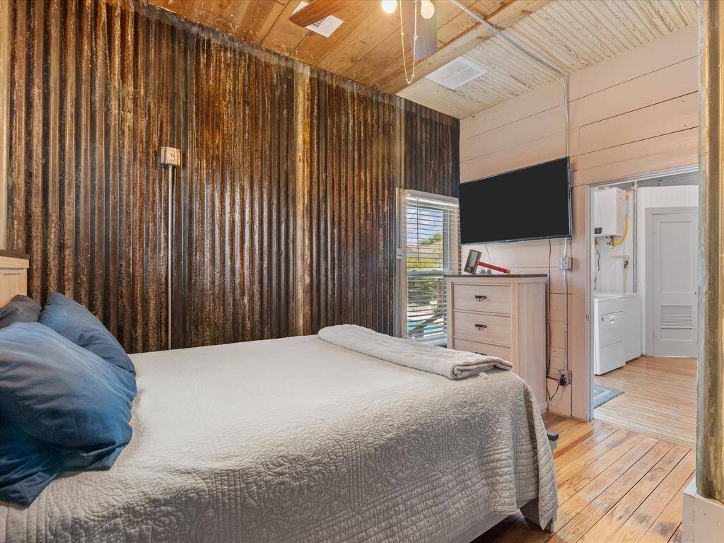 Cozy bedroom with rustic charm, featuring a corrugated metal accent wall and wooden ceiling. The space includes a comfortable bed, wall-mounted TV, and access to a laundry area. Ideal for those seeking a blend of industrial and homey aesthetics.