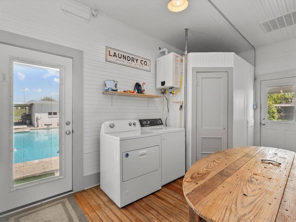 This photo features a bright, functional laundry room with a washer and dryer, a rustic wood table, and a view of a pool through a glass door, creating a seamless indoor-outdoor connection.