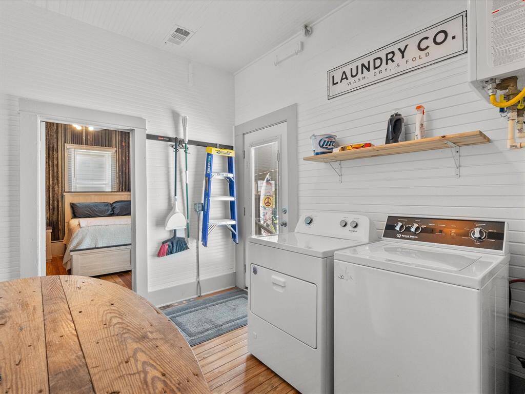 This photo shows a bright, functional laundry room with a washer and dryer, wooden flooring, and a shelf for supplies. There's a door leading outside and a glimpse into a cozy adjacent bedroom.