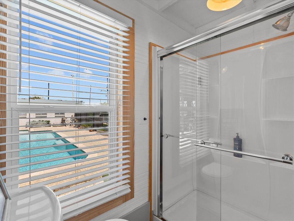 This photo shows a bright bathroom with a glass shower and a window featuring blinds. The window provides a view of a spacious outdoor pool area, adding a sense of luxury and relaxation to the home.