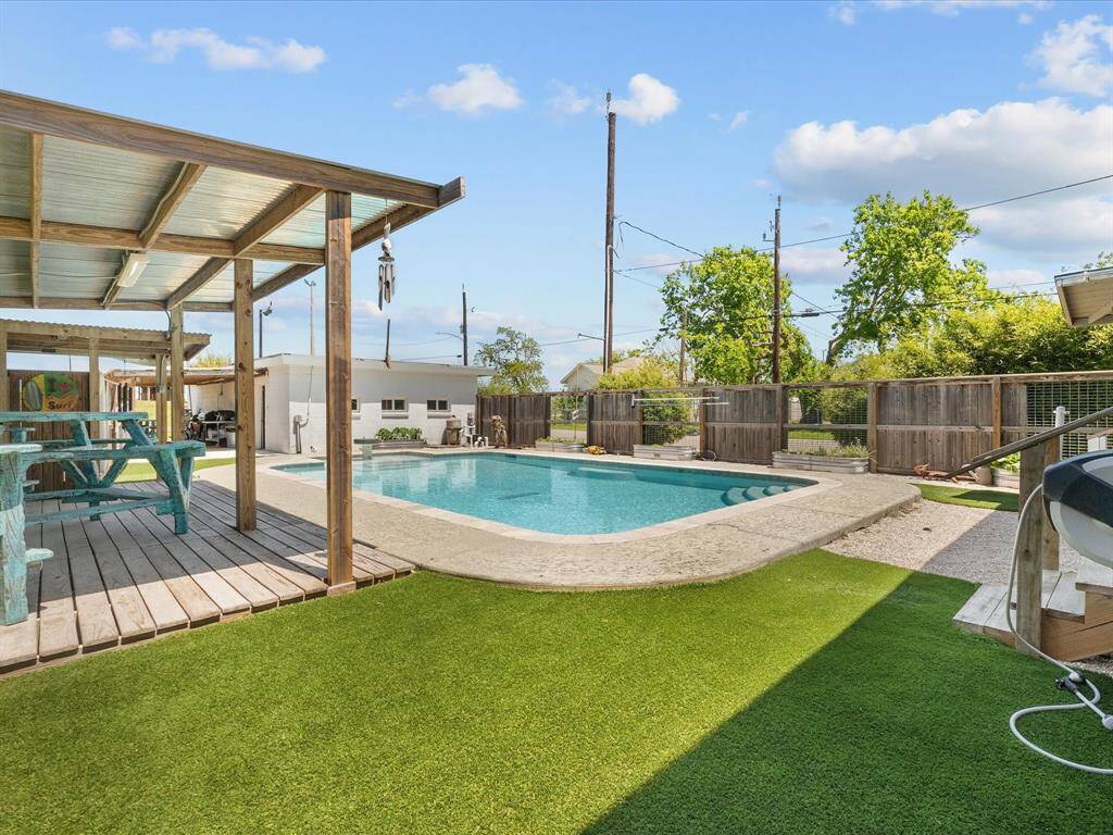 This backyard features a refreshing pool surrounded by a spacious deck and artificial grass. A covered patio area with a picnic table offers a great space for outdoor dining and relaxation.