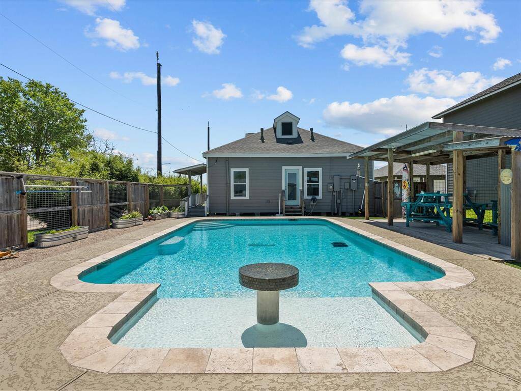 This photo features a charming backyard with a spacious in-ground pool, surrounded by a textured patio. A covered seating area with picnic tables is adjacent to the pool, perfect for outdoor gatherings. The house, painted in a neutral tone, provides a cozy backdrop.