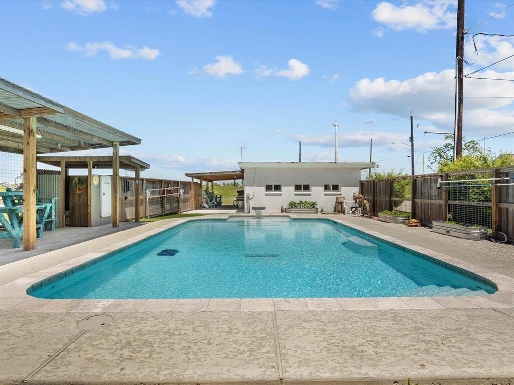 This photo features a spacious backyard with a large in-ground swimming pool, surrounded by a concrete deck. There's a covered patio area with seating, and the space is enclosed by a tall wooden fence, offering privacy. Perfect for outdoor relaxation and entertaining.