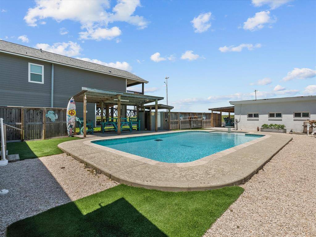 This photo features a spacious backyard with a well-maintained swimming pool, surrounded by a mix of artificial grass and gravel. A wooden pergola with seating provides a shaded area, perfect for relaxation or entertaining. The setting is private, charming, and ideal for enjoying outdoor activities.