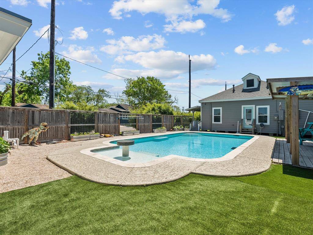This backyard features a spacious in-ground pool with a textured concrete deck and artificial grass surrounding it. A cozy gray house with a small patio is in the background, and the area is enclosed by a wooden privacy fence. Perfect for outdoor relaxation and entertaining.