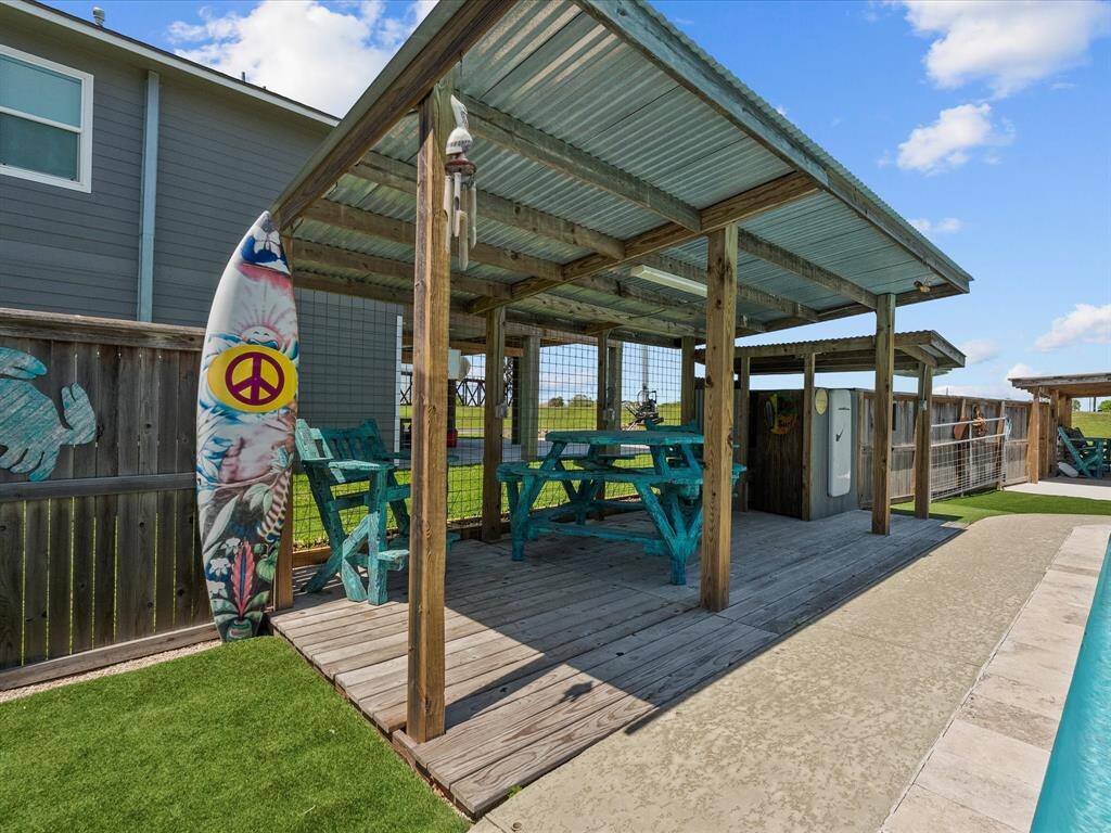 This photo showcases a cozy, covered outdoor seating area with turquoise picnic tables, set on a wooden deck. It features a surfboard with a peace sign, decorative elements, and is adjacent to a pool, creating a relaxed, beachy vibe perfect for entertaining or lounging.