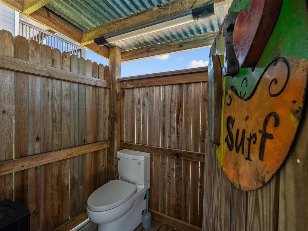 This photo shows an outdoor bathroom with a wooden privacy fence and a corrugated metal roof. It features a toilet and a surf-themed sign, creating a casual, beachy vibe.