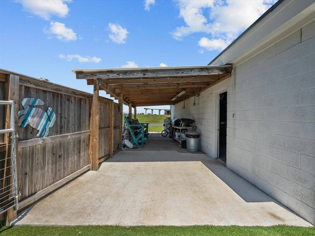 This photo shows a covered outdoor space with a wooden pergola attached to a building. There is a concrete floor, a grill, and some seating, ideal for outdoor activities. A wooden fence provides partial privacy, and there's a view of greenery beyond.