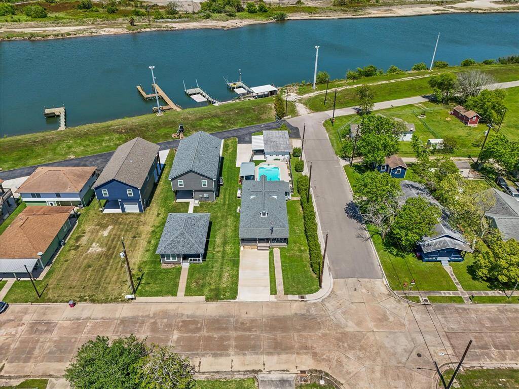 This aerial view shows a charming neighborhood with houses along a waterfront. The homes have well-maintained lawns and one features a backyard pool. A serene waterway with private docks runs behind the properties, offering scenic views and recreational opportunities.