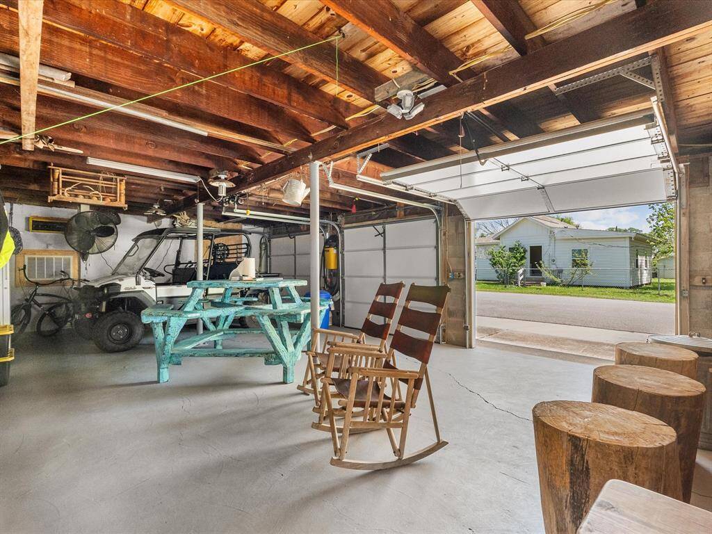Spacious garage with exposed wooden beams, concrete floor, and a large open door leading to the street. It features a teal picnic table, rocking chairs, and ample storage space for vehicles and equipment.