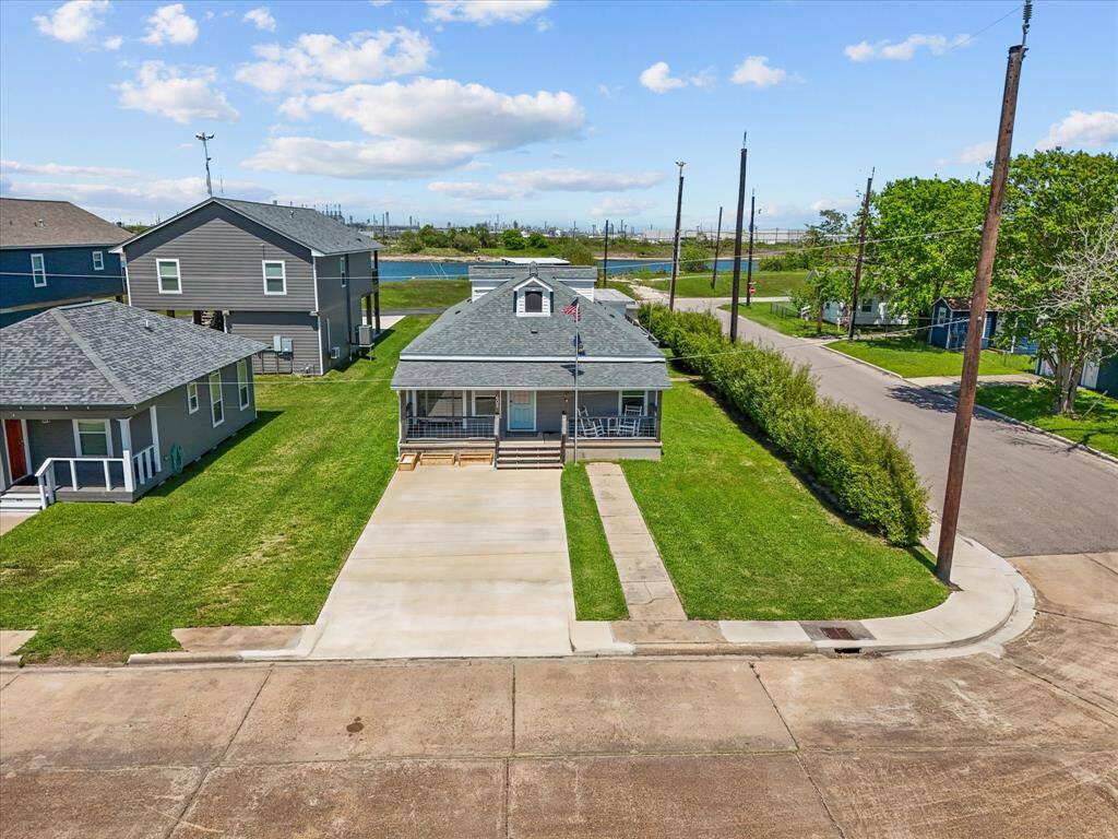 Charming single-story home with a gray roof and inviting front porch. It features a spacious driveway, well-maintained lawn, and is situated on a corner lot in a quiet neighborhood. The surrounding area includes greenery and a nearby body of water, offering a serene setting.