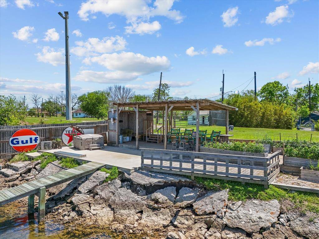 This photo shows a charming waterfront area featuring a wooden deck with seating, a rustic pergola, and vintage Gulf and Texas signs. The space is ideal for outdoor gatherings, overlooking a rocky shore with a small dock extending into the water. Lush greenery surrounds the property, enhancing its scenic appeal.