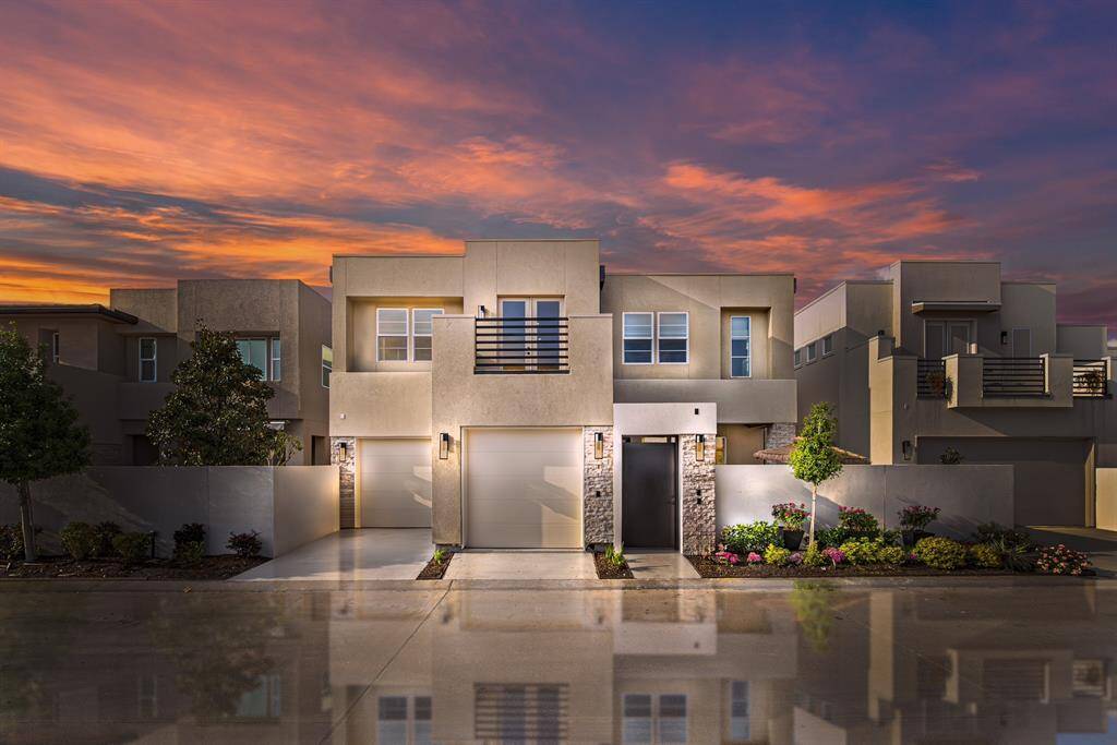 This modern two-story home features a sleek facade with clean lines, two garages, and landscaped surroundings. It’s set against a stunning sunset, highlighting its contemporary design and inviting curb appeal.