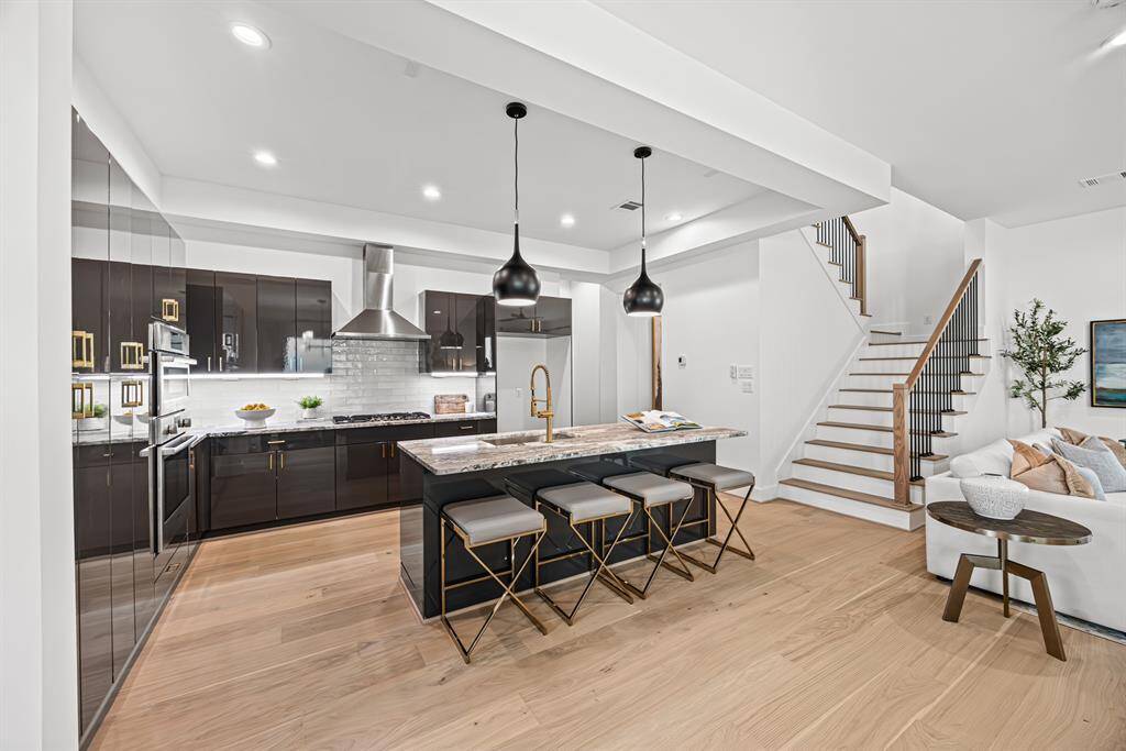 This modern kitchen features sleek dark cabinetry, a large island with seating, and stylish pendant lighting. It opens to a bright living area with a staircase, creating a spacious and inviting layout.