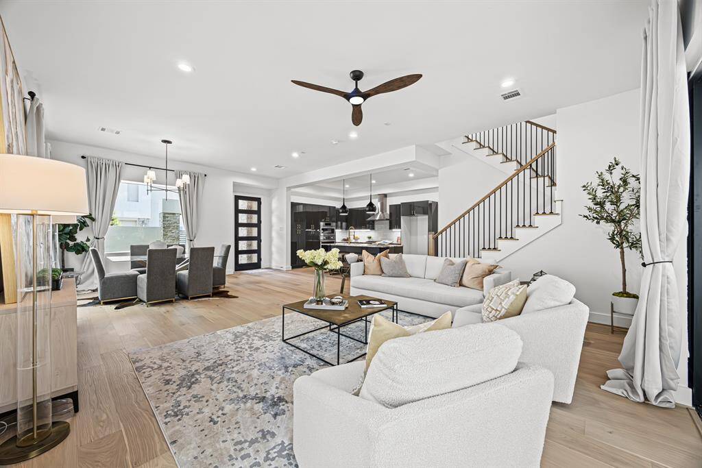 This modern, open-concept living area features a spacious layout with a cozy seating arrangement and stylish decor. The room is bright with large windows, a ceiling fan, and light wood floors. Adjacent is a dining area and a sleek kitchen, complemented by a contemporary staircase, creating an inviting and functional space.