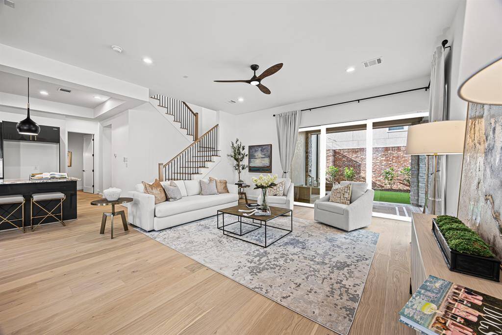 This spacious living area features modern design elements, including light wood flooring and a neutral color palette. It includes a cozy seating arrangement, a sleek kitchen with an island and bar stools, and large windows leading to a small outdoor space. A staircase adds architectural interest, and the room is well-lit with recessed lighting and a ceiling fan.