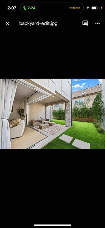 This photo showcases a modern backyard with a seamless indoor-outdoor living space. The area features a cozy seating arrangement on a wooden deck, large sliding doors, and a well-maintained lawn with stepping stones, framed by tall greenery for privacy.