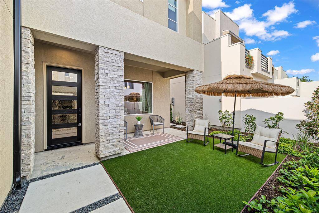 This photo showcases a modern patio area with a combination of stone and stucco finishes. It features a cozy seating area with chairs and a small table under a straw umbrella, set on a well-maintained artificial grass lawn. The space is bordered by neat landscaping and offers a stylish, low-maintenance outdoor retreat.