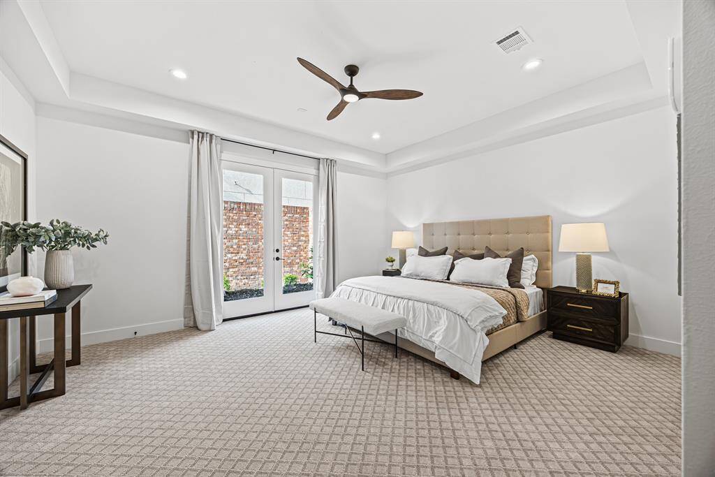 This bedroom features a modern, spacious design with a neutral color palette. It includes a large bed, elegant nightstands, soft carpeting, and ample natural light from double doors leading outside. The ceiling fan and recessed lighting add a touch of sophistication.