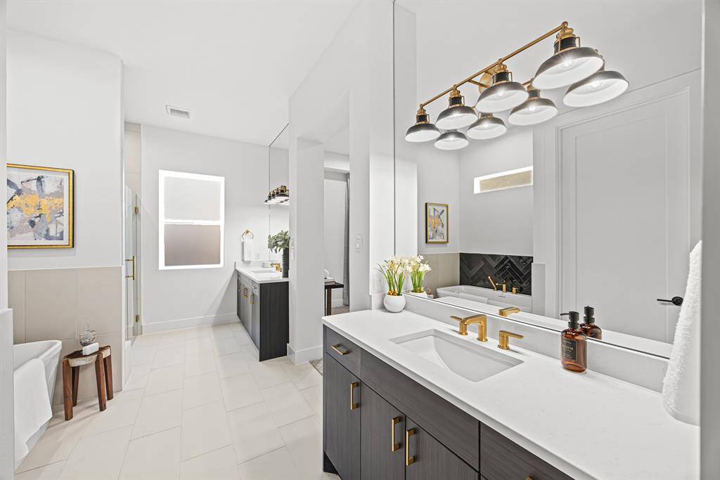 This bathroom features a modern design with sleek white countertops and gold fixtures. It includes dual sinks, a large mirror, and a spacious layout with ample natural light. The decor is minimalistic, with elegant lighting and subtle artwork.