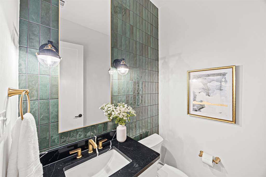 This bathroom features elegant green tiles, a large mirror, and gold accents. It has a modern sink with black countertop, stylish light fixtures, and tasteful wall art, creating a sophisticated and inviting space.