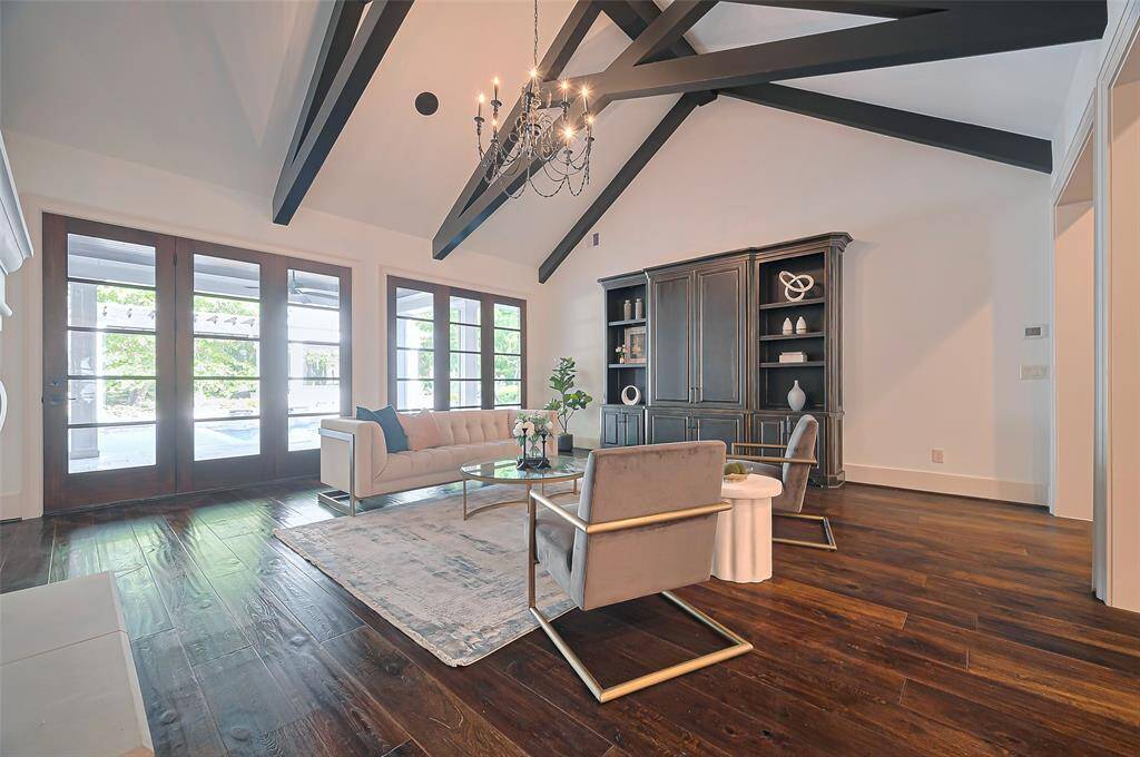 Large living room features high ceilings with exposed beams, a chic chandelier, and large French doors that let in plenty of natural light. The space tastefully holds large pieces of furniture or even a baby grand piano. Stylish entertainment cabinets, all set on rich hardwood floors.