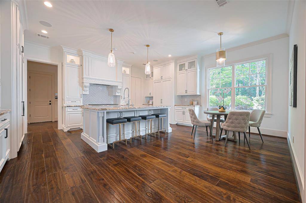 Gourmet Chef's Kitchen features elegant white custom cabinetry, a large island with seating, and modern pendant lighting. It has rich hardwood floors and a cozy dining area by a large window that offers plenty of natural light.