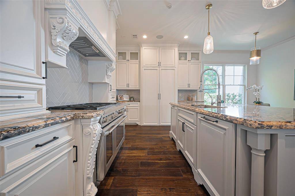 Sleek herringbone-patterned backsplash, and a high-end stainless-steel oven. The granite countertops and dark wood flooring add warmth. Cabinets reach to the high ceiling.